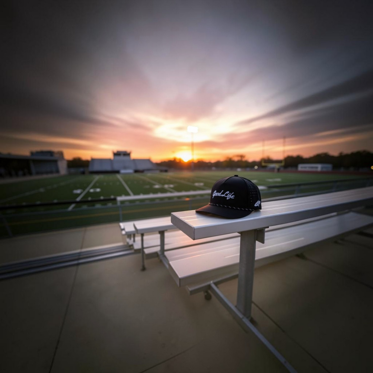 A black snapback cap with white Good Life script embroidery sits on white stadium bleachers overlooking a football field at sunset.