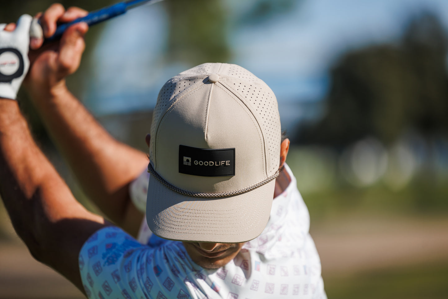 Person wearing a cap with 'Goodlife' branding, holding a golf club outdoors.