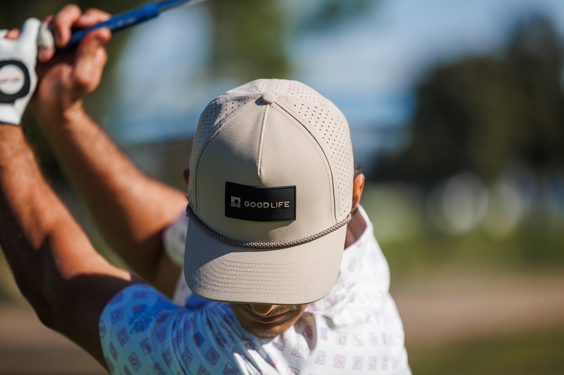 Person wearing a cap with 'Goodlife' branding, holding a golf club outdoors.