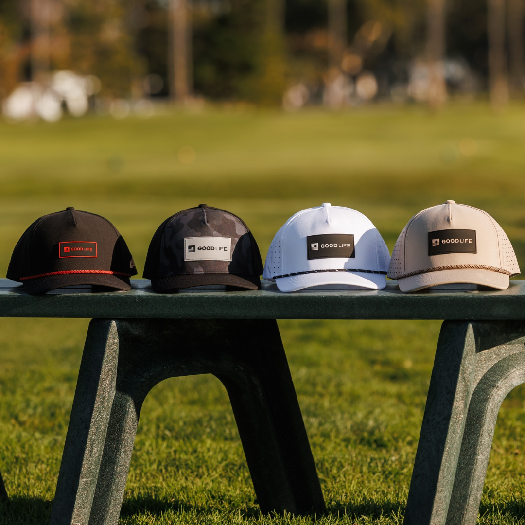 Four Good Life Golf performance rope hats in black, black camo, white, and khaki lined up on a green bench at a golf course