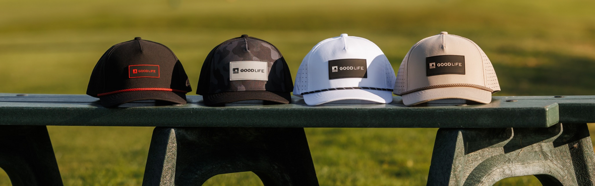 Four Good Life Golf performance rope hats in black, black camo, white, and khaki lined up on a green bench at a golf course