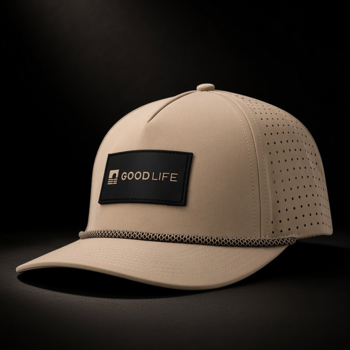 A beige snapback cap with perforated side panels, a black rectangular front patch reading GOOD LIFE, and a textured trim along the brim, photographed on a dark studio background.