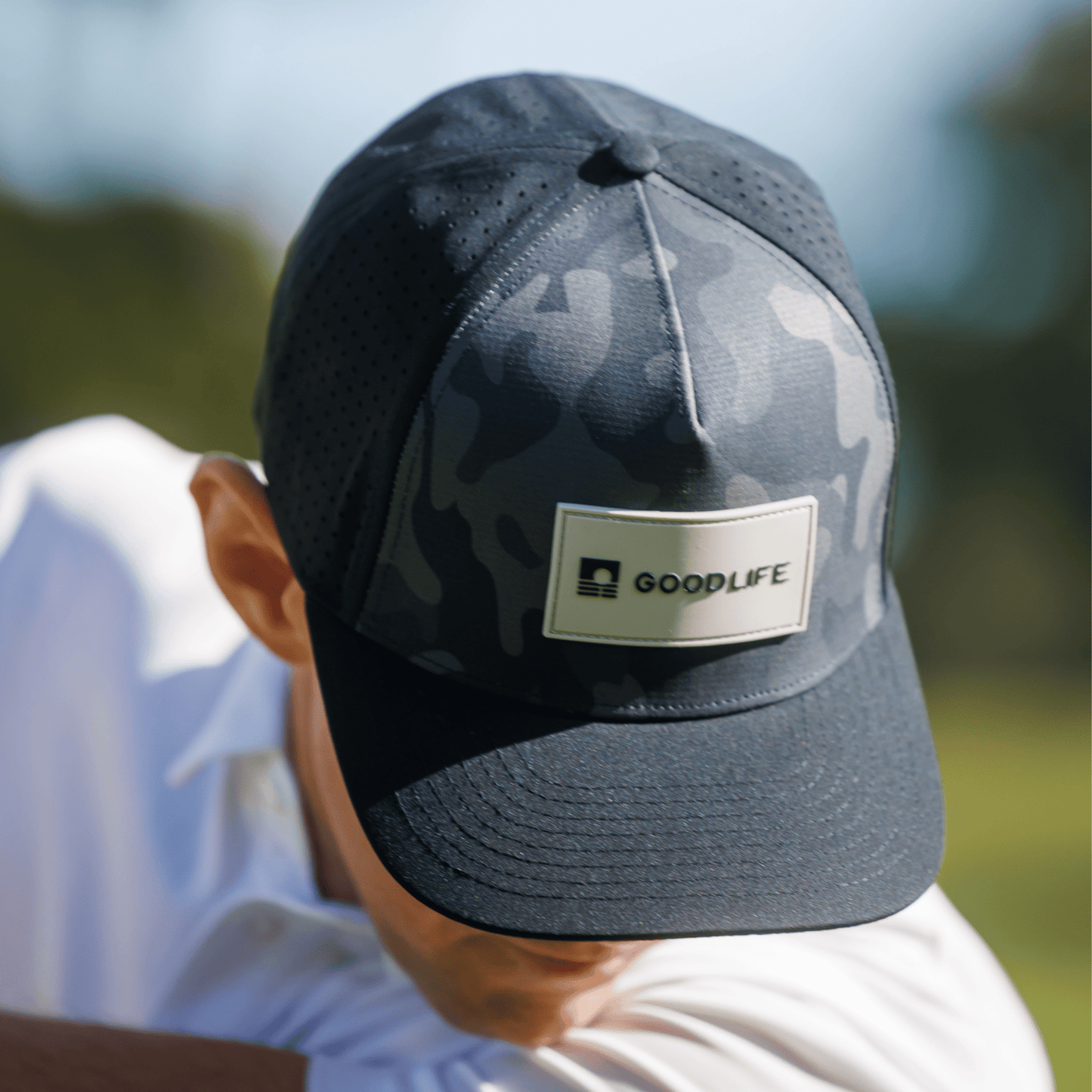 Person wearing a Good Life Golf Performance Black Camo Snapback with gray and black PVC patch water and sweat resistant perforated sides outdoors