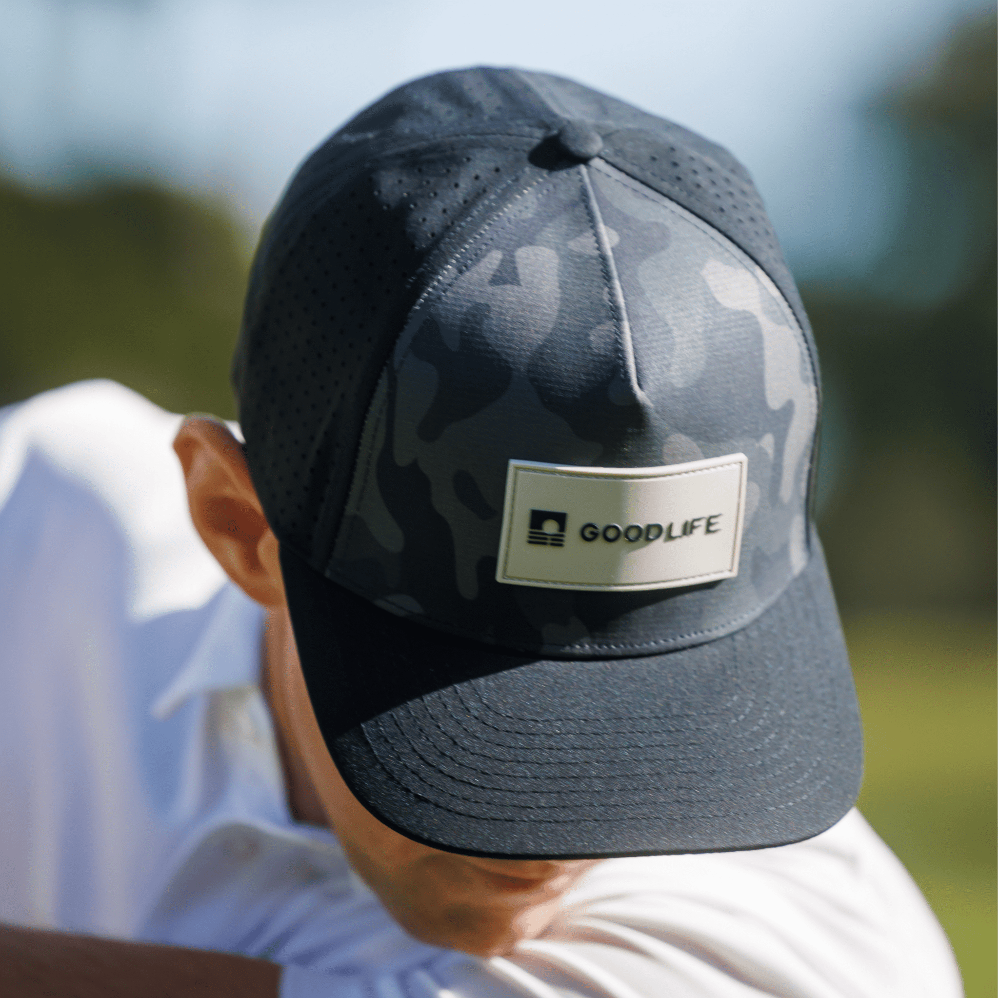 Person wearing a Good Life Golf Performance Black Camo Snapback with gray and black PVC patch water and sweat resistant perforated sides outdoors