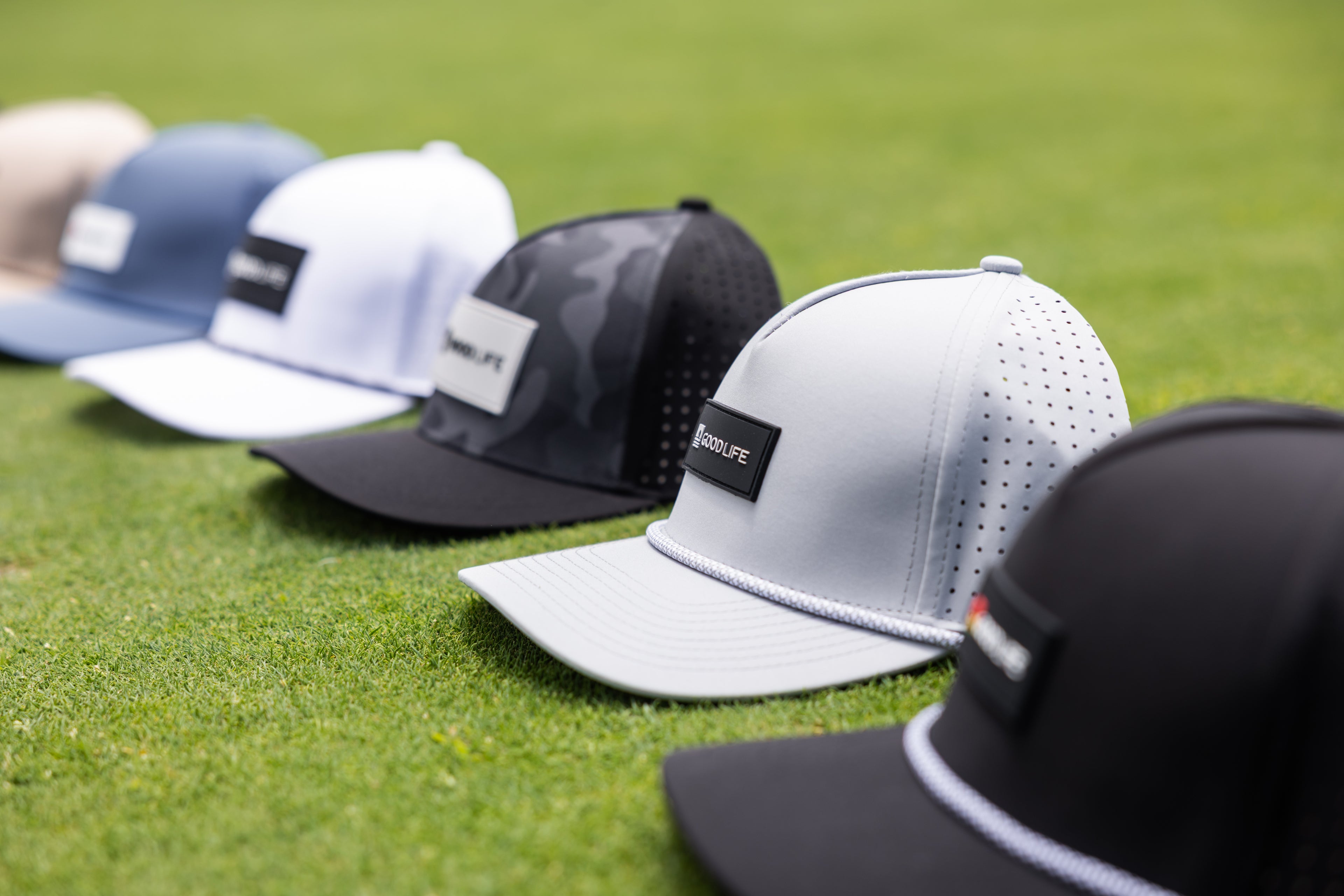 Row of Good Life Golf snapback hats on a grassy background