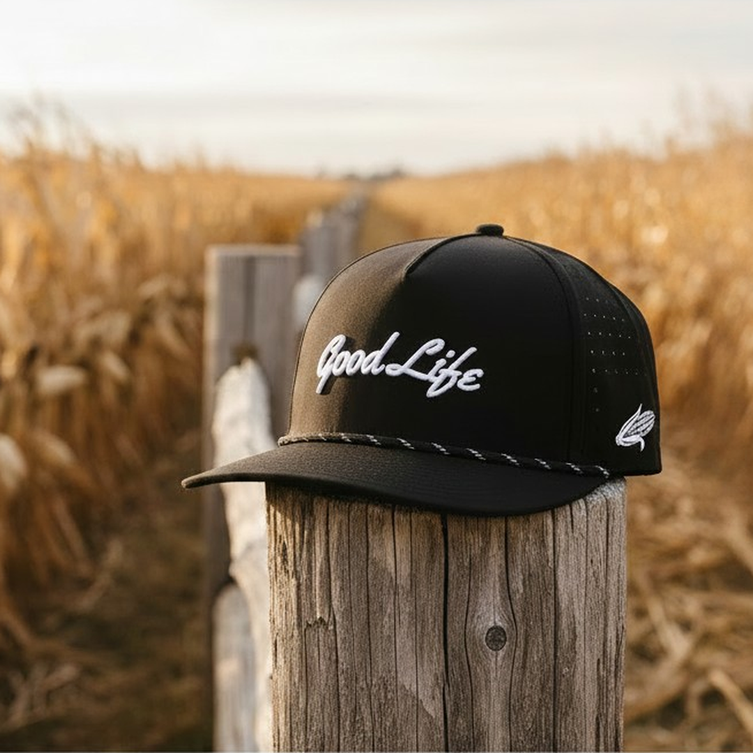 A black snapback cap with white embroidered 'Good Life' script on the front, resting on a weathered wooden post in a blurred golden field.
